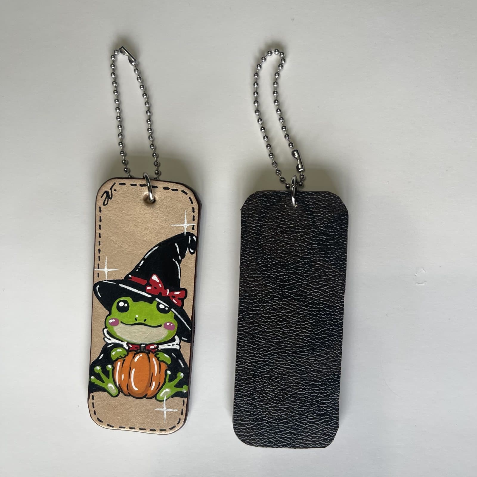 Leather charm painted with a smiling frog in a black witch hat and cape holding a pumpkin, beside its dark leather back
