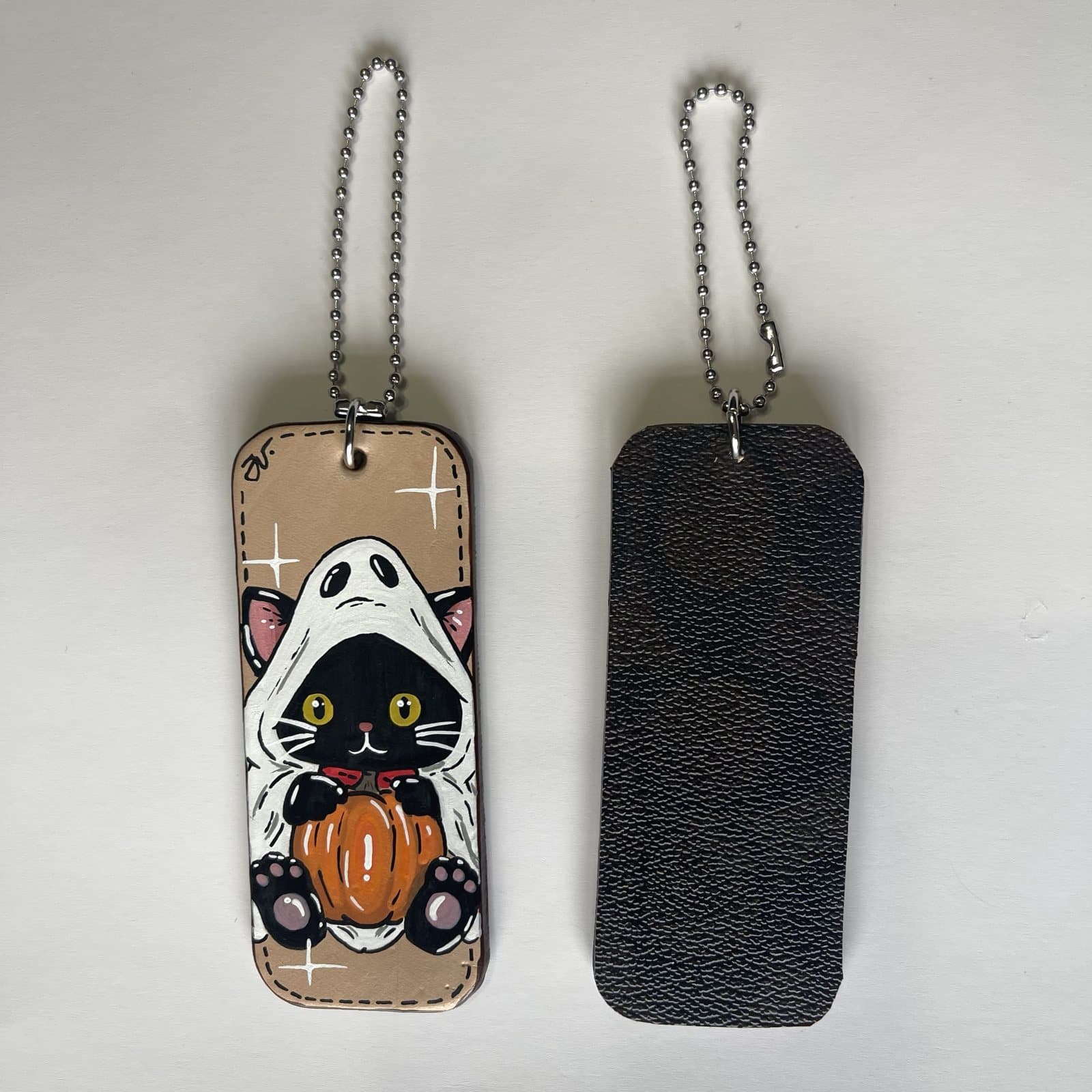 Leather charm painted with a yellow-eyed black kitten in a ghost costume holding a pumpkin, beside its dark leather back