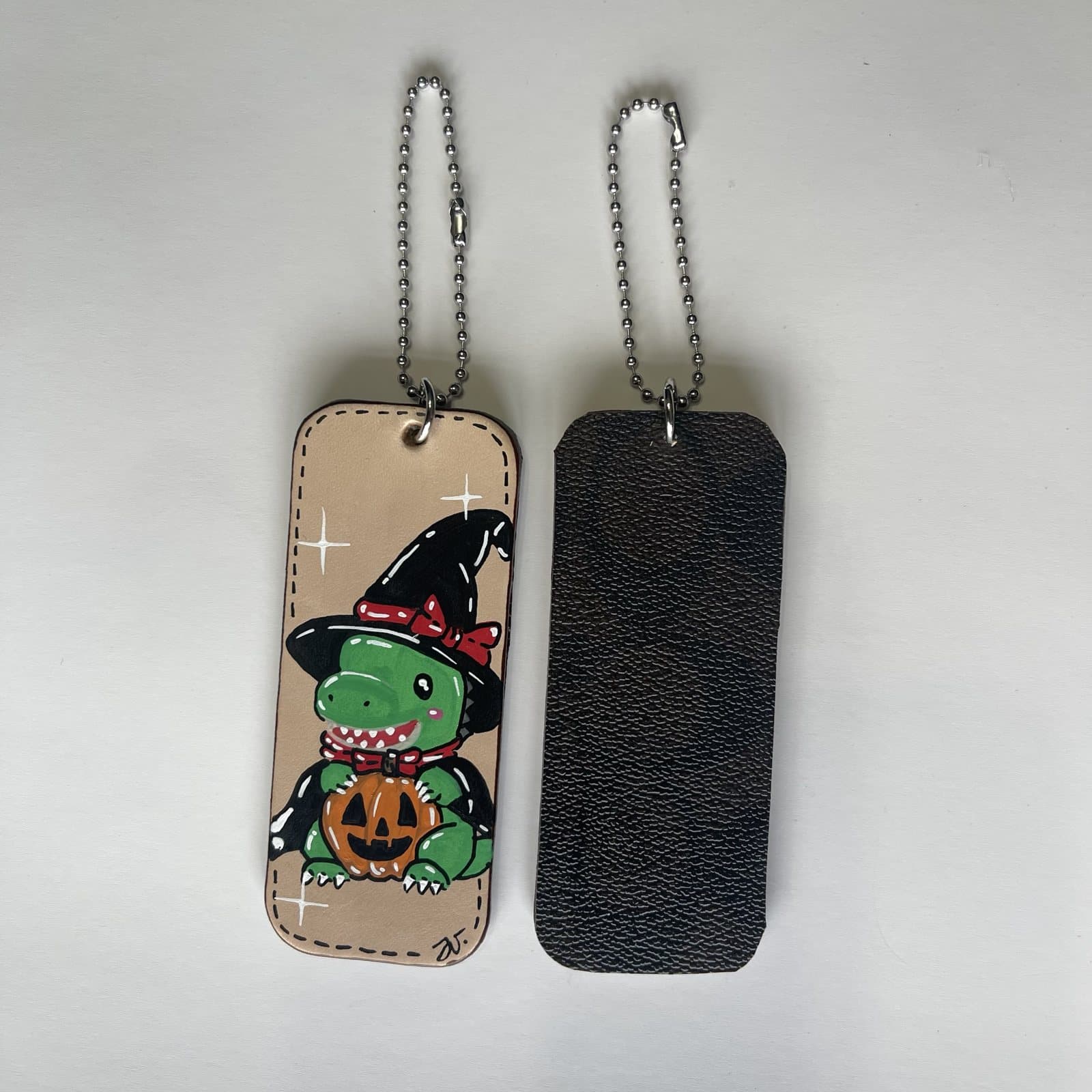 Leather charm painted with a green dino in a black witch hat with a red bow, holding a jack-o'-lantern, beside its dark leather back