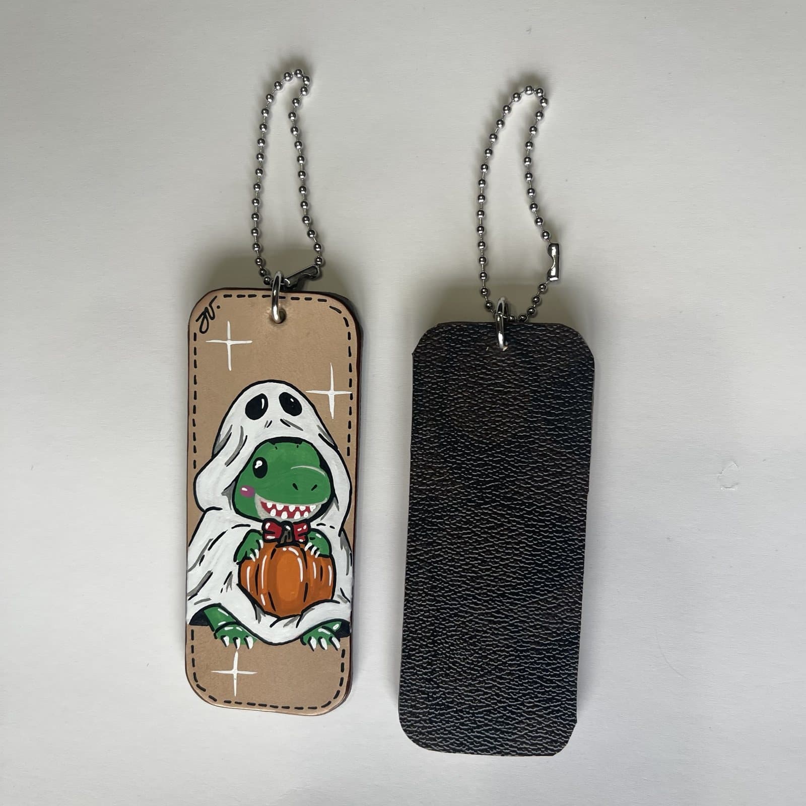 Leather charm painted with a green dino peeking out of a white ghost sheet, holding a pumpkin, beside its dark leather back