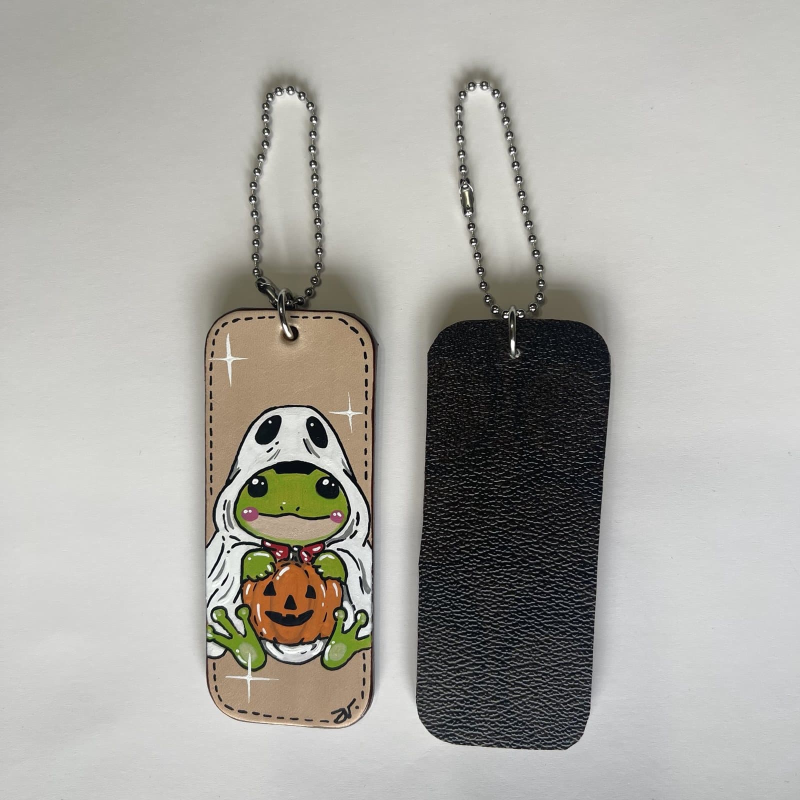 Leather charm painted with a frog in a ghost hood holding a smiling jack-o'-lantern, beside its dark leather back